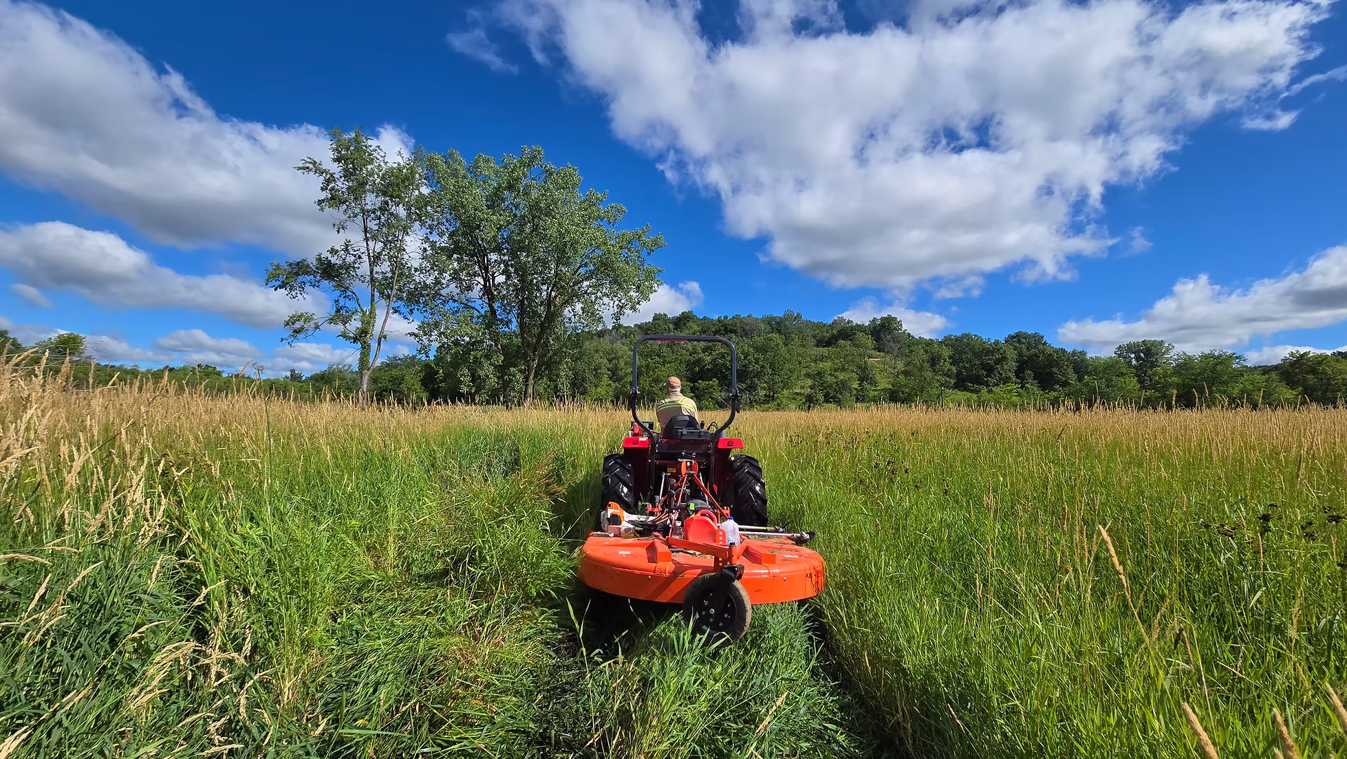 A person drives a lawn mower through tall prairie grass.